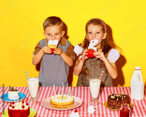 Happy, cheerfully adorable kids, siblings celebrating birthday, eating cakes against yellow background. Concept of childhood, birthday celebration, family, fun, food