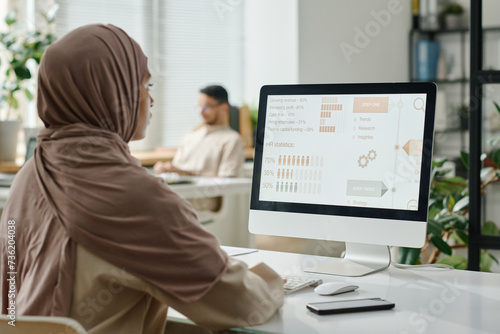Young Muslim businesswoman in hijab sitting by desk in front of computer screen with statistic data and analyzing it against male coworker photo