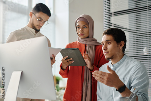 Two young Muslim employees using tablets while standing by their colleague looking at computer screen and explaining online data