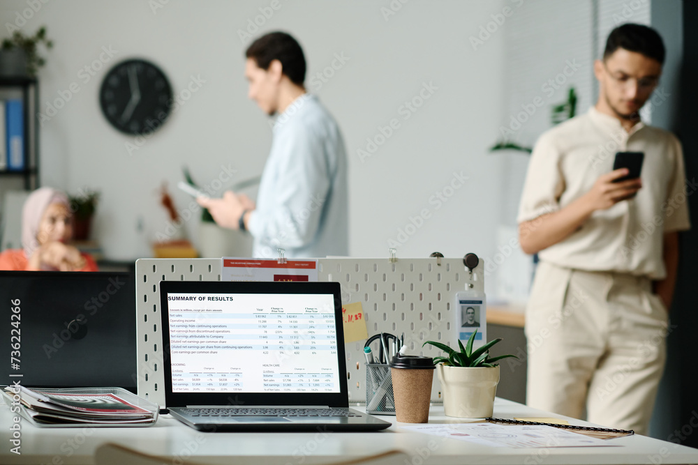 Laptop with summary of results on screen standing on desk which is workplace of analyst or broker against young employees