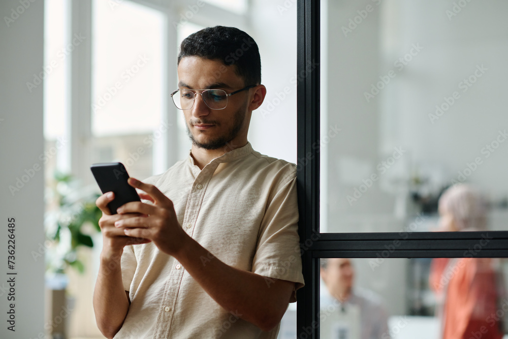 Young Muslim office manager or economist looking at smartphone screen while standing in coworking space against communicating colleagues Young Muslim office manager or economist looking at smartphone screen while standing in coworking space against communicating colleagues