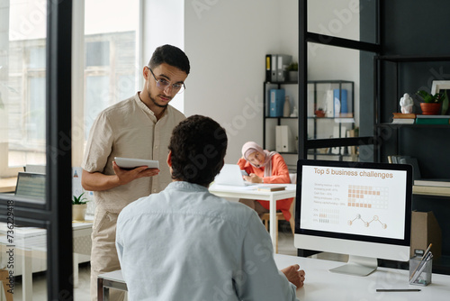 Two young male managers looking at one another during discussion while one of them sitting in front of desktop computer with data