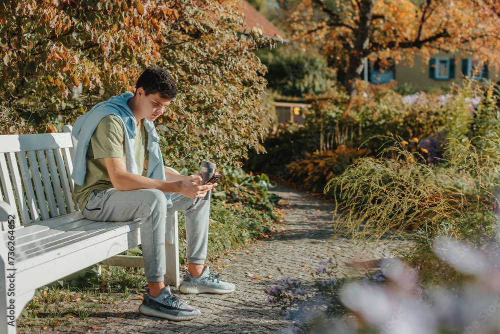 Fototapeta premium A Teenager Sits On A Bench In The Autumn Park Drinks Coffee From A Thermo Mug And Looks Into A Phone. Portrait Of Handsome Cheerful Guy Sitting On Bench Fresh Air Using Device Browsing Media Smm