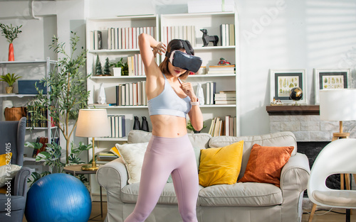 Asian healthy sportive woman wearing sportswear, VR glasses, playing game, standing in indoor living room at cozy home, doing body combat, boxing, exercising. Technology, Sport, Lifestyle Concept.