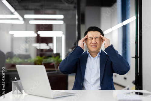 A man has a headache at the workplace inside the office, an Asian businessman in a business suit is sick, has a severe migraine, the boss is sitting at a desk with a laptop.