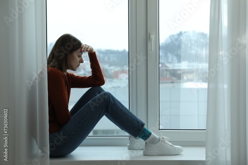 Sad young woman sitting on windowsill near window at home, space for text Free Download Sad young woman sitting on windowsill near window at home, space for text - GFXLoad