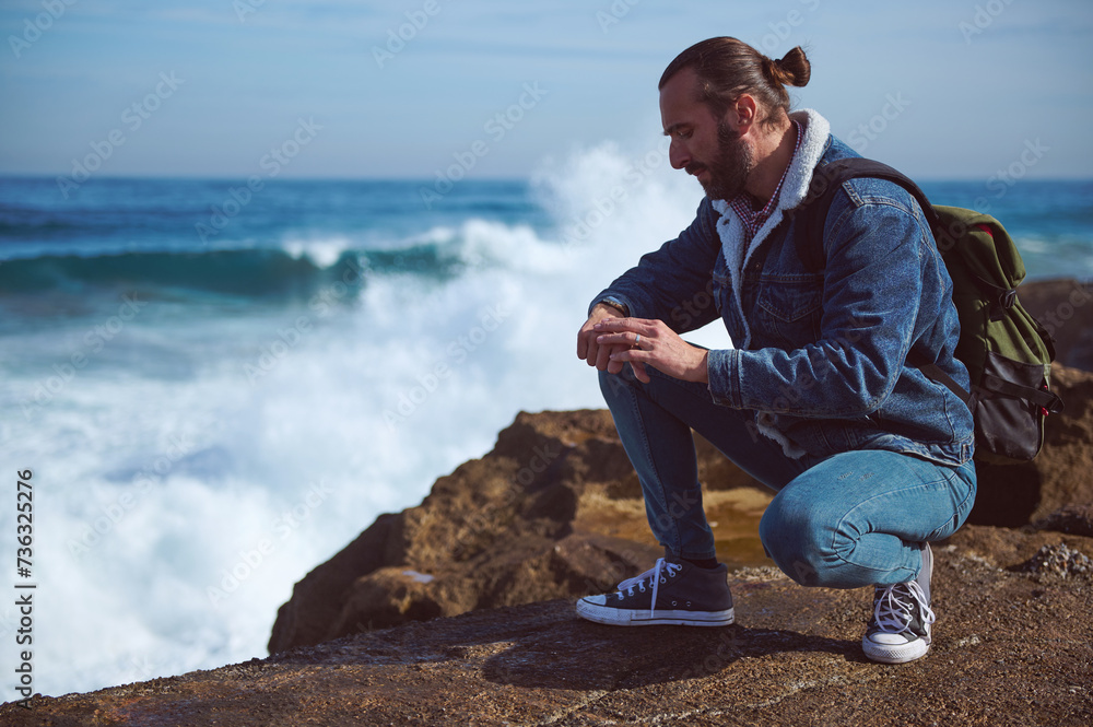 Handsome young male tourist checking his wristwatch, relaxing on the rocky cliff by sea with crushing waves. People. Travel. Discovering the beautiful wild nature. Active tourism and lifestyle concept