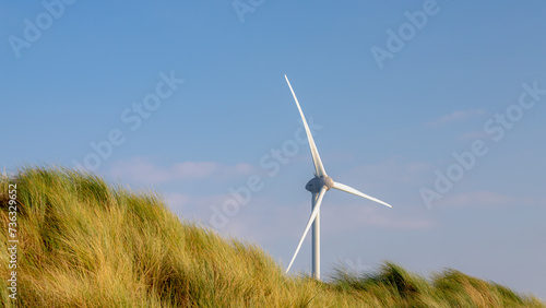 Fototapeta Naklejka Na Ścianę i Meble -  Clean energy concept with sand dune and european marram beach grass, White wind turbine with blue blue clear sky, Alternatively referred to as a wind energy converter, Countryside of the Netherlands.