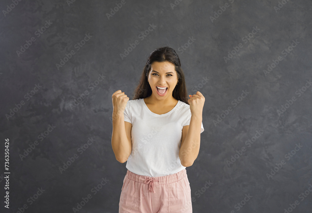 Portrait of a happy emotional woman laughing and rejoicing with clenched fists celebrating victory. Studio shot of a excited and joyful woman in casual clothes on a gray background. Concept of luck.