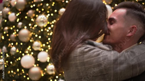 Romantic young couple spinning and kissing in front of large decorated Christmas tree. Loving girlfriend and boyfriend rubbing noses and embracing in shopping mall. Winter holidays concept. photo