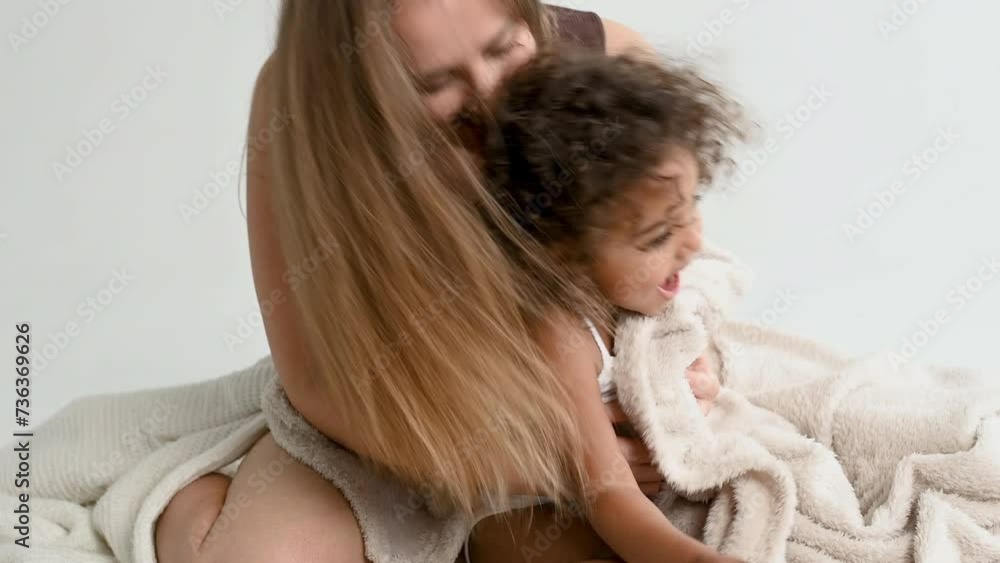 Interracial family dressed in combidresses is having fun on white background sitting on the floor and plays hide and seek with white cozy plaid. European mother and 2 years black african daugh Interracial family dressed in combidresses is having fun on white background sitting on the floor and plays hide and seek with white cozy plaid. European mother and 2 years black african daugh