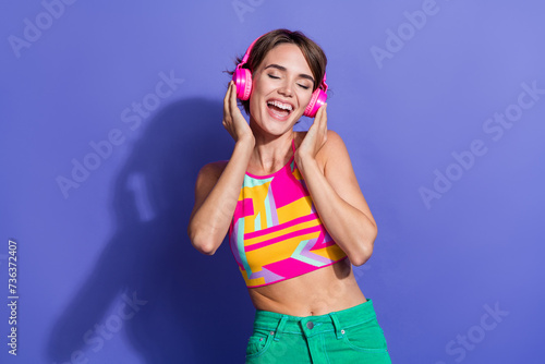 Portrait of youngster hipster woman in crop top singlet touching her bright pink headphones enjoy bass isolated on violet color background
