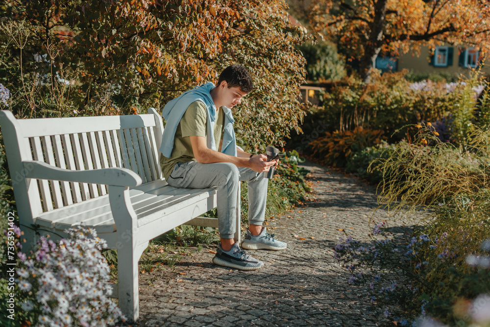 Fototapeta premium A Teenager Sits On A Bench In The Autumn Park Drinks Coffee From A Thermo Mug And Looks Into A Phone. Portrait Of Handsome Cheerful Guy Sitting On Bench Fresh Air Using Device Browsing Media Smm