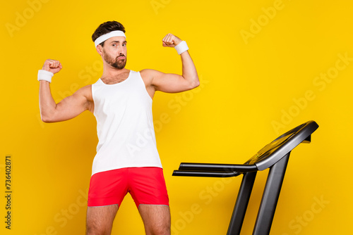 Portrait of confident powerful person treadmill equipment flexing biceps isolated on yellow color background