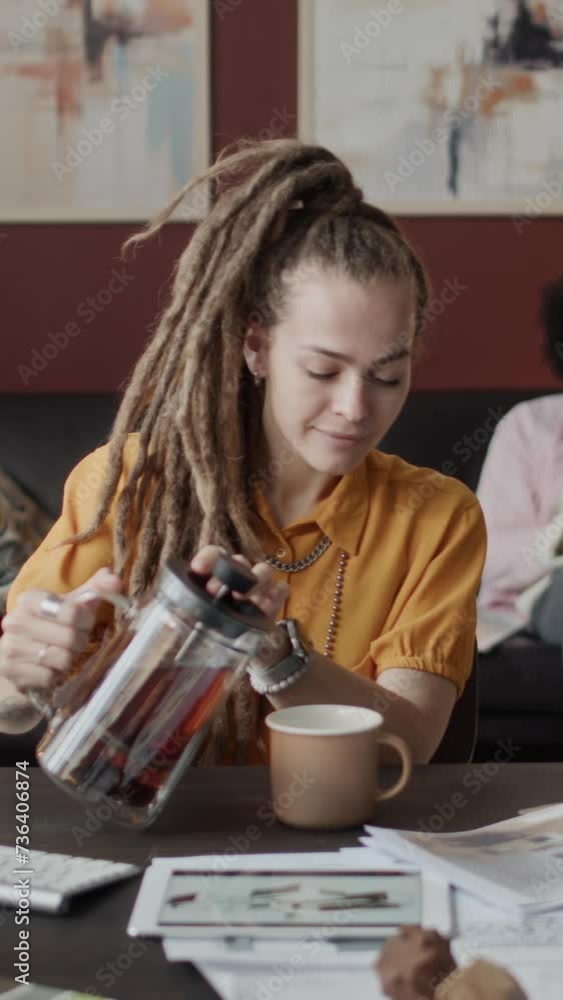 Vertical medium shot of young Caucasian female interior designer pouring and drinking calming hot tea and stretching her neck while sitting at desk at home office working on house design project Vertical medium shot of young Caucasian female interior designer pouring and drinking calming hot tea and stretching her neck while sitting at desk at home office working on house design project