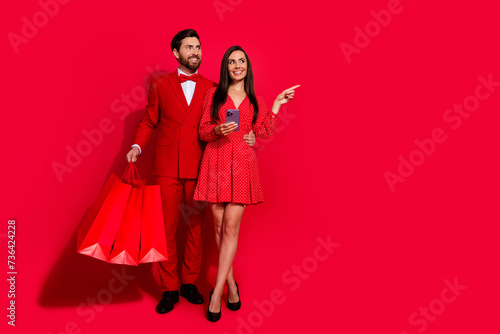 Full length portrait of two beautiful lovers embrace store bags smart phone indicate finger empty space isolated on red color background
