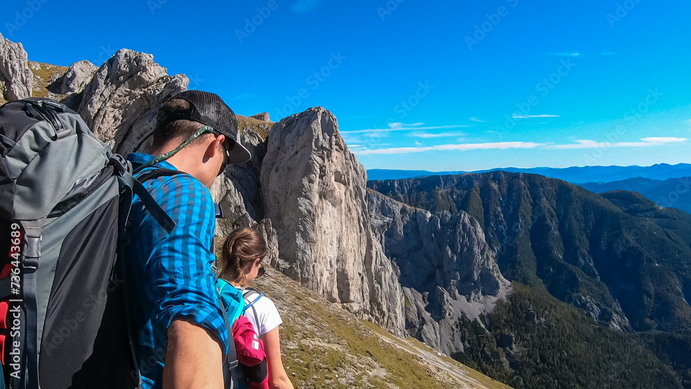 Naklejka premium Hiker couple on idyllic hiking trail on alpine meadow with scenic view of majestic Hochschwab mountain range, Styria, Austria. Wanderlust in remote Austrian Alps. Sense of escapism, peace, reflection