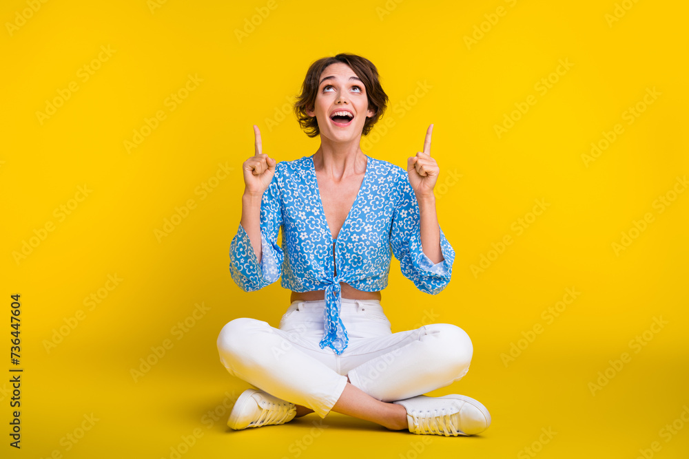 Full size photo of positive woman wear blue top sit on floor look directing at logo empty space isolated on yellow color background Full size photo of positive woman wear blue top sit on floor look directing at logo empty space isolated on yellow color background