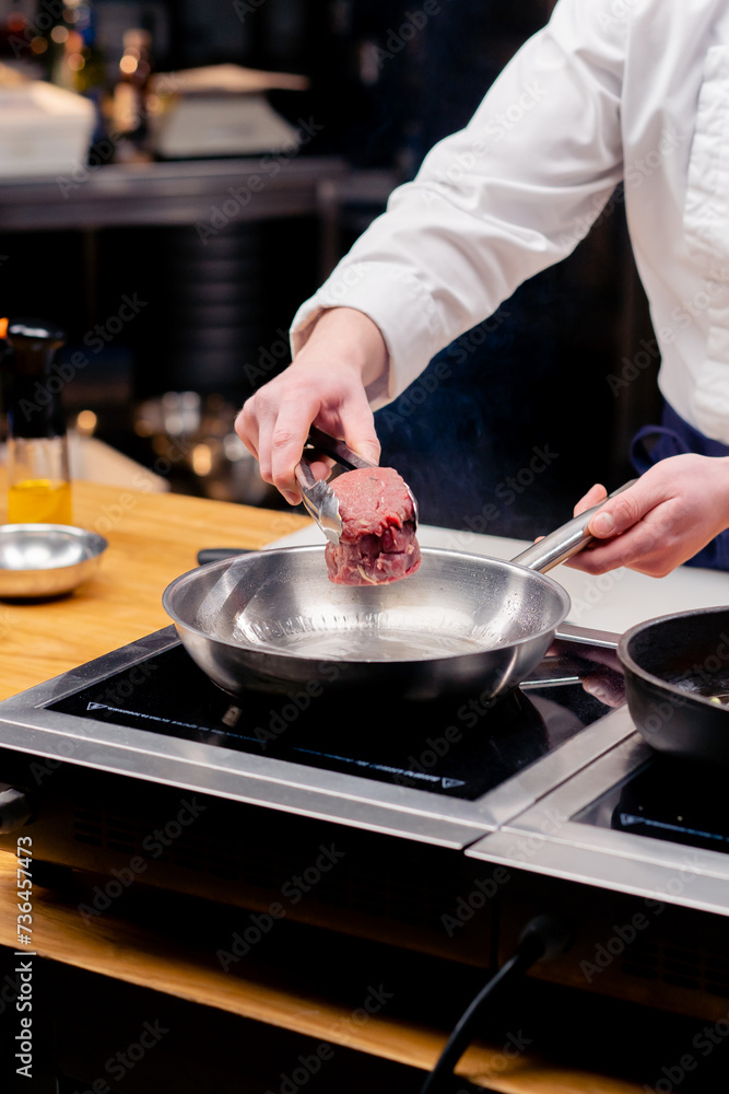 Fototapeta premium close-up of a chef's hand placing a beef steak on frying pan with tongs