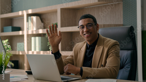 Happy African American man working with laptop computer at office remote job business commerce professional businessman looking at camera smile waving hand hello hi friendly sign greeting online meet