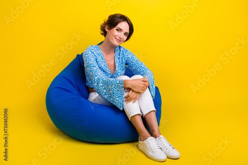 Full size photo of cheerful gorgeous woman wear blue top sitting on bean bag embrace her knees isolated on yellow color background photo