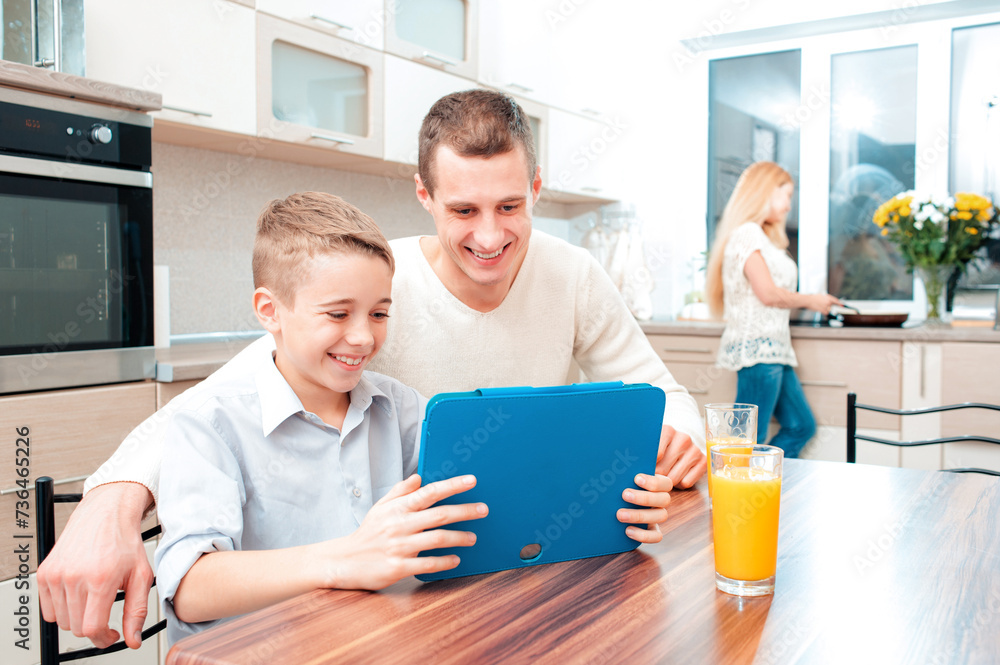 Obraz premium Happy together. Boy and his father using a digital tablet and laughing while mother cooking dinner at the background.