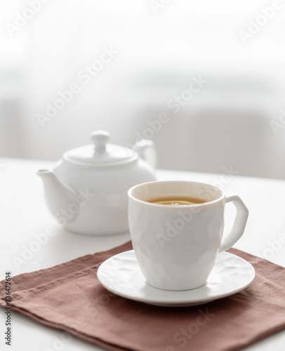 A cup of tea with lemon and a teapot on a white table against the background of a kitchen window.