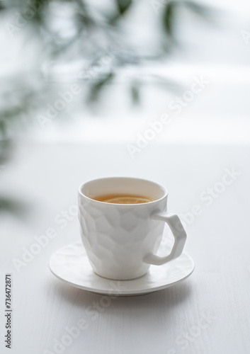 A cup of tea with lemon and green branch on a white table against the background of a kitchen window.