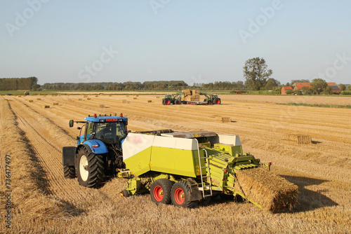 a rural landscape with a tractor and a big baler in front at a straw field and tractors with front loaders in the background photo