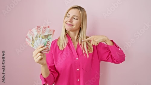 Cheerful young blonde woman proudly displaying new zealand dollars, pointing with optimistic smile, over isolated pink background