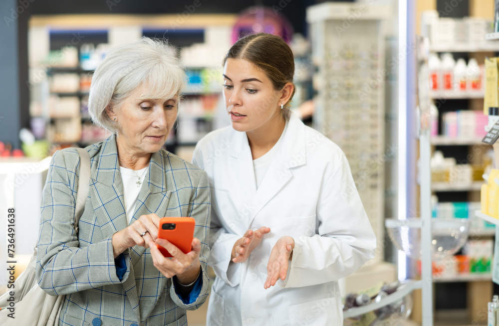 Concerned old female customer asking young chemist about product reflected on her phone in large pharmacy