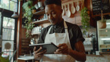 A male cafe worker is using a tablet with a smile inside a well-lit coffee shop, surrounded by various coffee shop elements.