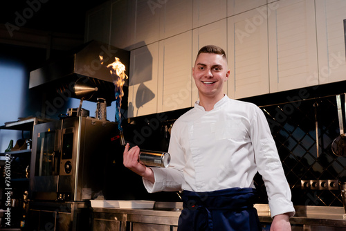 A chef in a white jacket in a professional kitchen stands with working burner in his hand and smiles