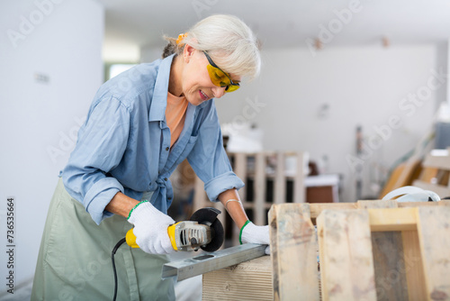 Focused mature woman in protective glasses cutting metal profile using grinding machine, renovating house with her own hands