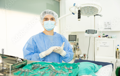 Professional young veterinarian in sterile coat  cap  mask and gloves preparing to conduct surgical operation at veterinary clinic  standing by operating table  confidently looking into camera..