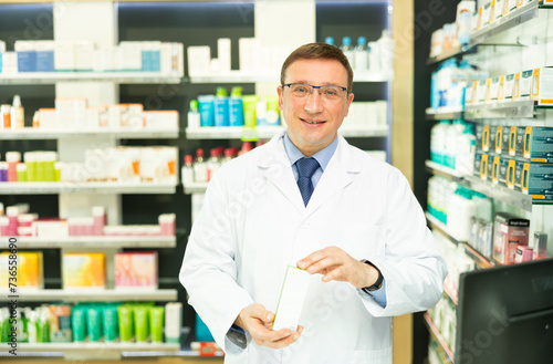 Portrait of man druggist in lab coat standing in drugstore with drug package © JackF