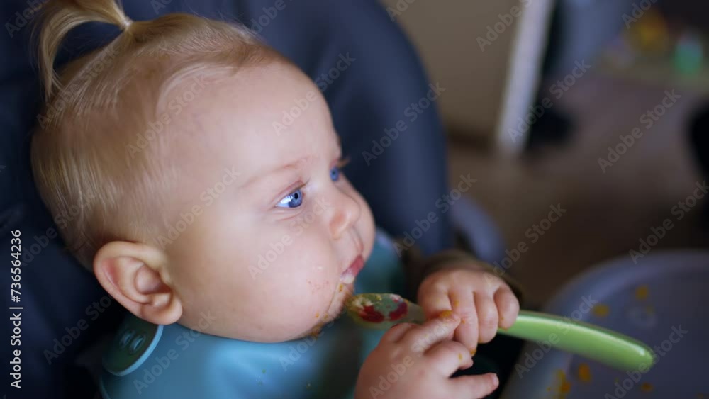 Face of a blond blue-eyed infant with a bib. Little kid is chewing a spoon during meals. Close up. Face of a blond blue-eyed infant with a bib. Little kid is chewing a spoon during meals. Close up.