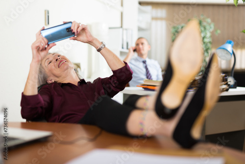 Senior woman office worker sitting in workplace with her legs on table and using smartphone. Her colleague talking on phone in background.