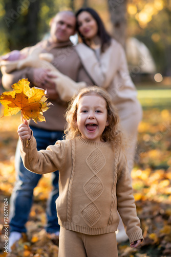 Happy young family with two children dressed in beige colors stands in the park.  The eldest daughter holds leaves in her hands