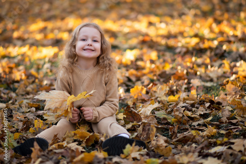 Little girl in a beige knitted suit sits in a park in autumn leaves and rejoices