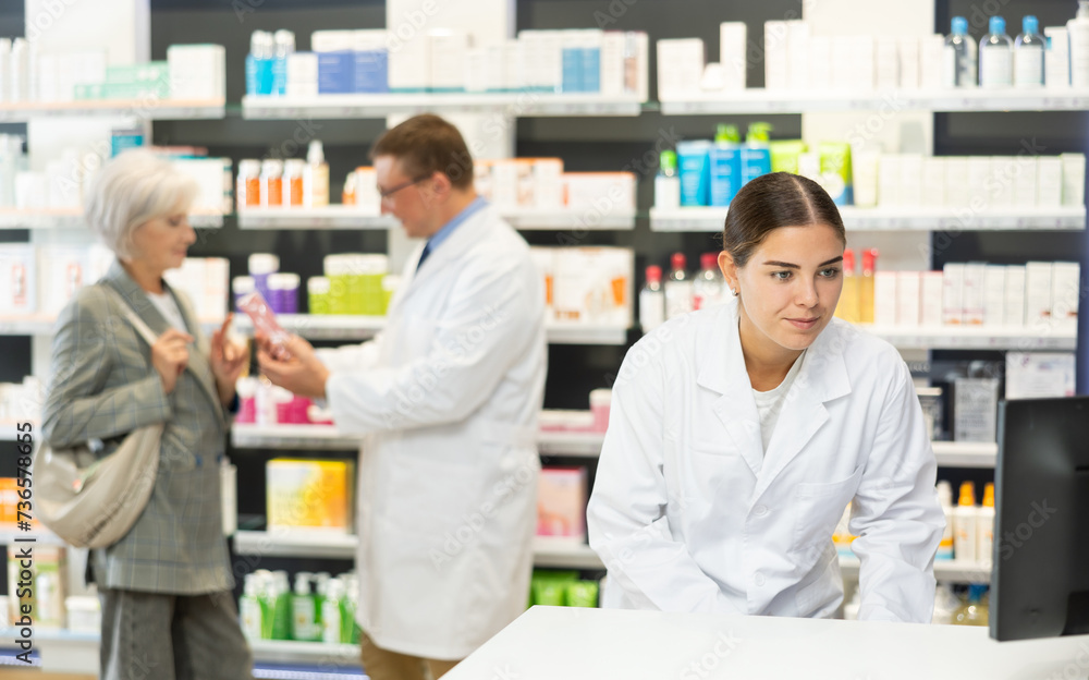 Girl pharmacist in pharmacy works with computer, searches among nomenclature of drugs, specifies price and availability of medicine - obrazy, fototapety, plakaty 