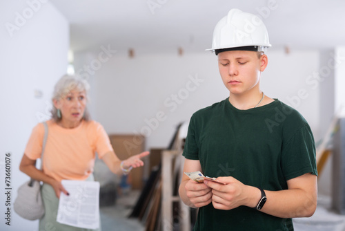 Portrait of a disappointed fifteen-year-old teenager with money in his hands, working at a construction site indoors, to ..whom a disgruntled mature female client underpaid part of his salary photo