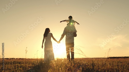 Father and wife strolling with daughter atop shoulders and silhouettes blending into sky in evening. Silhouettes of father carrying daughter on shoulders with wife strolling to horizon at sunset photo