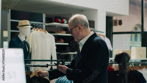 Trendy senior man shopping for new fashion collection at retail store, visiting modern clothing shop to check formal or casual items. Rich customer uses phone to look for products by code. Camera A. photo