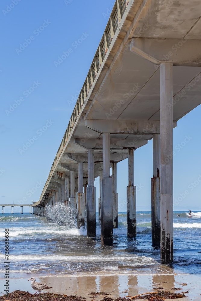Portion of Concrete Pier Crossing Frame From Right to Left, with Waves Crashing into Pylons and Distant Surfers, Shot from Underneath, San Diego, California, USA, vertical