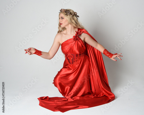 Full length portrait of beautiful blonde model dressed as ancient mythological fantasy goddess in flowing red silk toga gown, crown. Kneeling pose sitting on floor. isolated on white studio background