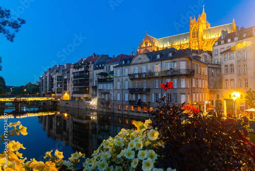 Scenic nighttime view of summer Metz cityscape overlooking illuminated majestic Gothic Roman Catholic Cathedral towering over residential buildings on waterfront along Moselle River, France
