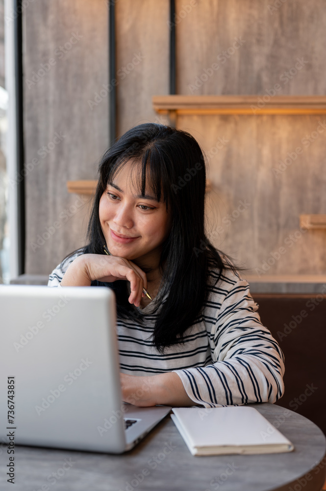 A happy Asian woman is working remotely at a cafe co-working space, looking at her laptop screen. A happy Asian woman is working remotely at a cafe co-working space, looking at her laptop screen.