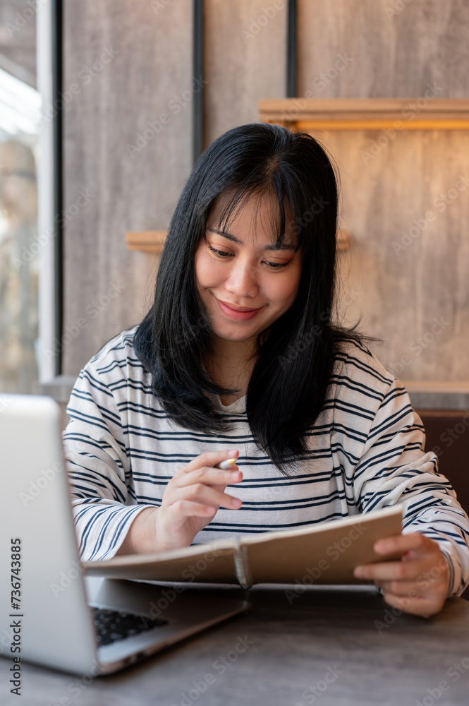 A happy Asian woman is enjoying listing her ideas in a book while working remotely at a coffee shop. A happy Asian woman is enjoying listing her ideas in a book while working remotely at a coffee shop.