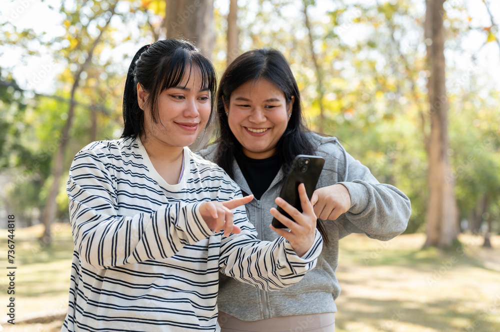 Two young Asian female best friends looking at a smartphone screen together while standing in a park Two young Asian female best friends looking at a smartphone screen together while standing in a park